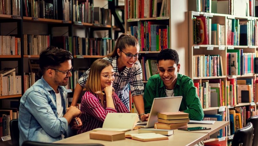 Vier Personen sitzen in einer Bibliothek an einem Tisch und arbeiten gemeinsam mit Laptop und Fachbüchern. Szene zeigt konzentriertes Recherchieren und Prüfen von Informationen.